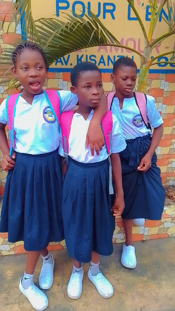 Trois écolières en uniforme avec leurs sacs à dos roses posant devant la pancarte de la fondation